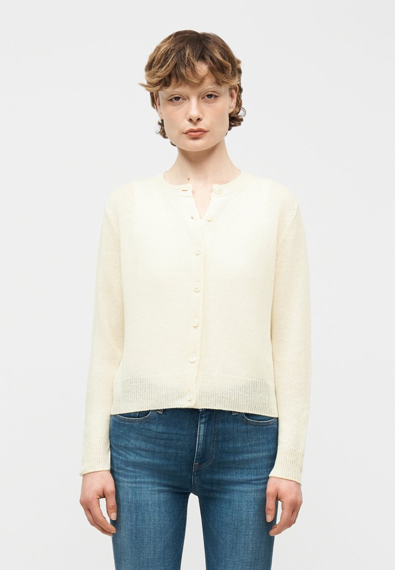 Νεαρό άτομο με κοντά σγουρά μαλλιά που φοράει κρεμ ζακέτα με κουμπιά και μπλε τζιν, στέκεται μπροστά σε απλό ανοιχτό φόντο.