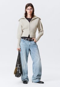 Beige gerippter Zip-Pullover, cropped, mit weiten Ärmeln. Hellblaue baggy Jeans. Schwarzer Gürtel und Lederschuhe, hält eine schwarze Tasche mit einem Pelzakzent.