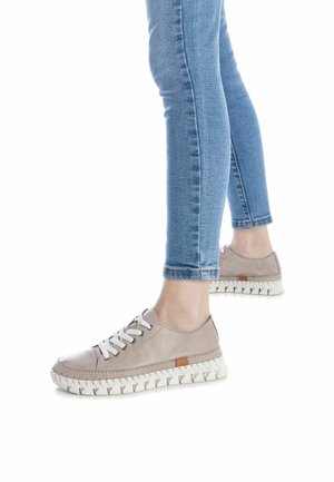 Zapatillas beige con cordones, con un upper de cuero texturizado, suela gruesa blanca y un acento marrón cerca del talón, combinadas con vaqueros azules ajustados.