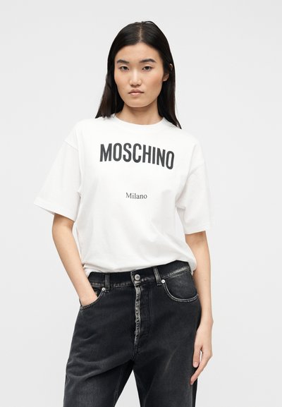 Νεαρή γυναίκα με μακριά σκούρα μαλλιά, φορώντας λευκό t-shirt Moschino Milano και μαύρα τζιν, στέκεται με το ένα χέρι στην τσέπη.