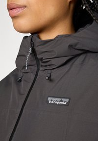 Close-up van een persoon die een donkergrijze Patagonia-jacket met capuchon en zichtbare rits draagt, met het merlogo op de borst.