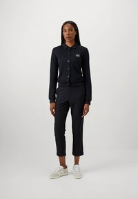 Πλεκτή μπλε ζακέτα με κούμπωμα με κουμπιά και λογότυπο, συνδυασμένη με μαύρο στενό παντελόνι και ανοιχτό γκρι αθλητικά παπούτσια. Απλός, δομημένος σχεδιασμός.