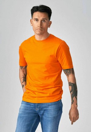 Mand med kort mørkt hår iført en lys orange T-shirt og blå jeans, der viser tatoveringer på begge underarme, stående foran en ensfarvet baggrund.