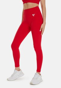 Leggings rouges taille haute en matière extensible, avec un détail de logo blanc sur la hanche et une texture lisse et élégante.