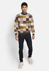 Mehrfarbiger Strickpullover mit einem Karomuster in Gelb, Braun, Marineblau und Creme, kombiniert mit dunklen Jeans und hellen Sneakers.