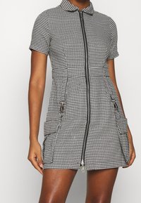 Lyhythihainen mustavalkoruudullinen mekko, jossa vetoketju edessä, kaksi etutaskua ja kaulus. Sileä tekstuuri ja räätälöity istuvuus.