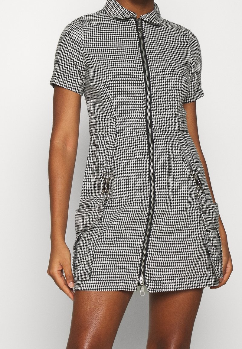 Lyhythihainen mustavalkoruudullinen mekko, jossa vetoketju edessä, kaksi etutaskua ja kaulus. Sileä tekstuuri ja räätälöity istuvuus.