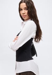 Νεαρή γυναίκα με μακριά καστανά μαλλιά, φορώντας λευκό πουκάμισο κάτω από μαύρο αμάνικο τοπ, στέκεται με σταυρωμένα τα χέρια μπροστά σε απλό φόντο.