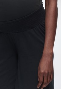 Σκούρα ελαστική ζώνη στη μέση μαύρων παντελονιών που φοριούνται από άτομο με σκουρόχρωμο δέρμα, δείχνοντας τη μέση, τους γοφούς και το δεξί χέρι να κρέμεται προς τα κάτω.