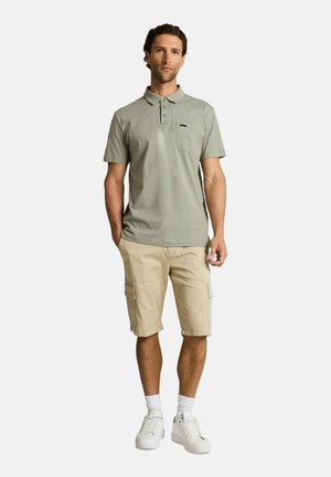 Hombre de pie con camisa polo verde salvia, pantalones cortos tipo cargo beige, calcetines blancos y zapatillas blancas frente a un fondo liso.