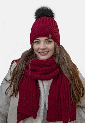 Beanie rosso a maglia con un pompon in pelo sintetico nero, abbinato a una sciarpa spessa e-texturizzata rossa. Entrambi presentano un design a coste e un materiale morbido.