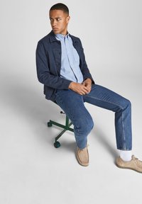 Chaqueta navy sobre una camisa de botones azul claro, combinada con jeans azules y zapatos beiges, sentado en un taburete verde ajustable.