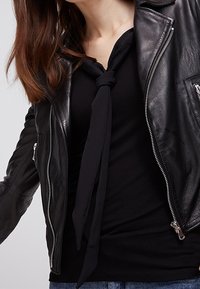 Svart läderjacka med silverfärgade dragkedjor, bärs över en svart blus med en knuten halsdetalj. Blå denimjeans synliga i nederkanten.