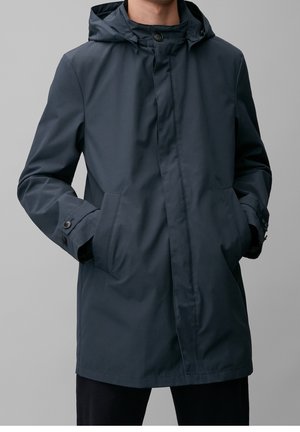 Mężczyzna ubrany w ciemnoszary płaszcz przeciwdeszczowy z kapturem, z napinanymi mankietami i rękami w kieszeniach na jednolitym szarym tle.