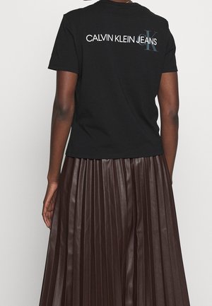 Czarna bawełniana koszulka z logo "CALVIN KLEIN JEANS" na plecach, zestawiona z długą, plisowaną, ciemnobrązową spódnicą.