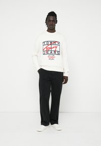 Weißer Pullover mit "Tommy Jeans"-Logo in Rot und Schwarz, kombiniert mit schwarzen Hosen und weißen Sneakern. Einfaches, lässiges Design.