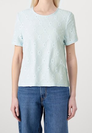 Vrouw draagt lichtblauw kort mouwig blouse met bloemrijk ajourpatroon en blauwe spijkerbroek, staand tegen een witte achtergrond.