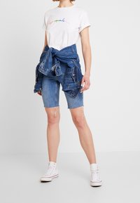 Bílý tričko s barevným textem, spárované s modrými denimovými šortkami. Džínsová bunda je uvázaná kolem pasu. Bílé tenisky doplňují vzhled.