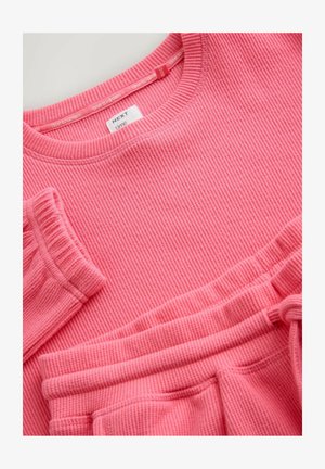 Roze geribbelde trui en bijpassende broek. Zachte textuur, ronde hals en elastische manchetten. Merklapje zichtbaar op de trui.