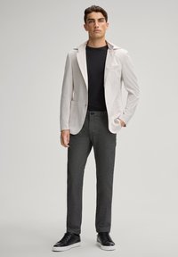 Blazer strutturato grigio chiaro sopra una camicia nera, abbinato a pantaloni scuri in tessuto testurizzato e sneakers nere. Presenta un colletto a scollo a V e tasche.
