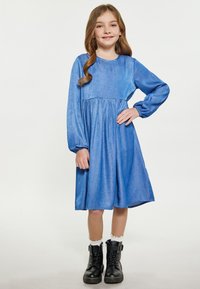 Blå denimkjole med lange pufferærmer, en rund halsudskæring og en samlet talje. Bæres med sorte snørebåndsstøvler og hvide ankelsokker.
