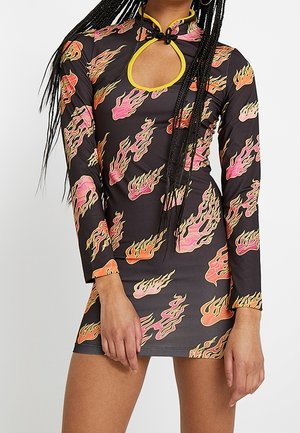 Femme portant une mini-robe noire à manches longues ornée de motifs de flammes roses et orange, avec un décolleté en trou de serrure à l'avant bordé de jaune.