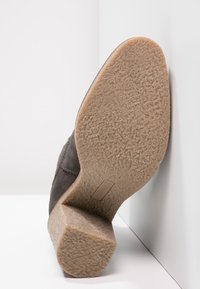 Tumma harmaa mokkanahkakenkä, jossa on kuvioitu kumipohja, kaareva muoto ja paksu kantapää, sijoitettuna valkoiselle pinnalle.