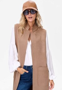 Beżowa bezrękawnik o teksturowanym wykończeniu, biała koszula z długim rękawem, dżinsy oraz skórzana czapka. Widoczne okrągłe przeciwsłoneczne okulary oraz detal w postaci kieszonki.