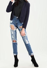 Navy kolor marynarka typu cropped nałożona na czarny i biały sweter z golfem, zestawiona z lekko niebieskimi spodniami dżinsowymi z dużymi przetarciami oraz czarnymi botkami na obcasie.