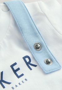 Bela tkanina srajca z svetlobluho naramnico, ki ima dve srebrni sponki in temno modro natisnjen napis "BAKER" spodaj.