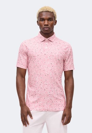 Man met krullend blond haar, gekleed in een nauwsluitend roze polo met korte mouwen en een lichtgekleurde broek, staand tegen een effen achtergrond.