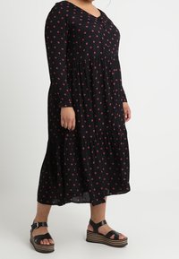 Robe midi noire avec un motif floral rouge, à manches longues, décolleté en V et jupe à volants. Associée à des sandales à plateforme noires.