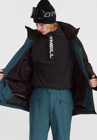Chaqueta de esquí verde azulado con forro negro, bolsillos con cremallera; combinada con un sudadera de forro polar negra y pantalones de esquí a juego, modelo lleva un gorro y gafas de esquí.