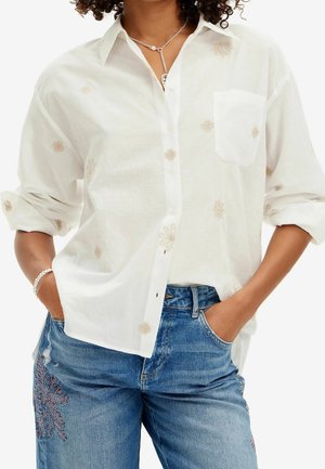 Femme portant une chemise blanche ample avec des motifs floraux légers et un jean bleu brodé, mains dans les poches et bracelet tressé au poignet.