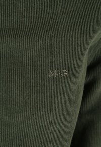 Olijfgroen geribd materiaal met subtiele textuur; voorzien van een klein geborduurd "MøG" logo in een bijpassende kleur. Geschikt voor casual kleding.