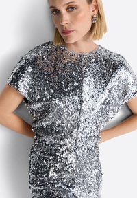 Blizganti sidabrinė suknelė, pagaminta iš blizgučių, su laisvu siluetu, trumpomis rankovėmis ir apvaliu iškirpimu. Jokios papildomos raštų ar detalių nematyti.