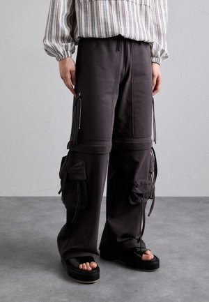 Graue Cargo-Hosen mit mehreren Taschen, verstellbaren Bindegürteln und Reißverschlüssen. Kombiniert mit schwarzen Slipper-Sandalen. Hat ein lässiges Design.