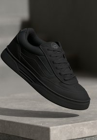 Zwarte sneaker met een gladde textuur, voorzien van een ronde neus, platte veters en zijaccenten. Het heeft een stevige rubberen zool en minimale stiksels.