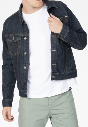 Veste en denim bleu foncé avec poches avant, coutures dorées et boutons argentés. Portée sur un t-shirt blanc et assortie à un pantalon vert clair.