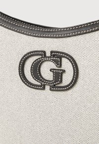 Torba na ramię z płótna, z czarnym skórzanym logo wyszywanym, beżowym korpusem o teksturowym wykończeniu oraz kontrastowym szwem wokół górnej krawędzi.