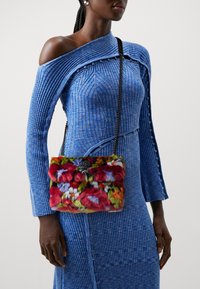 Bunte Fell-Umhängetasche mit einer plüschigen Textur. Sie verfügt über einen schwarzen Verschluss und einen Kettenriemen, kombiniert mit einem gerippten blauen Kleid.