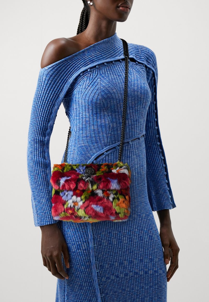 Bunte Fell-Umhängetasche mit einer plüschigen Textur. Sie verfügt über einen schwarzen Verschluss und einen Kettenriemen, kombiniert mit einem gerippten blauen Kleid.