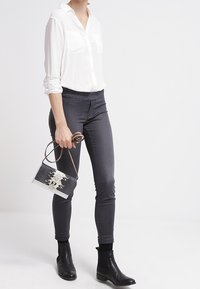 Camicia bianca con bottoni e due tasche sul petto, jeans skinny grigio scuro, stivali neri alla caviglia e una piccola borsa a tracolla con motivo.