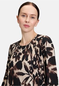 Frau mit heller Haut und blauen Augen, die eine langärmlige Bluse mit braunem und schwarzem Blumenmuster sowie plissiertem Stoff vor einem weißen Hintergrund trägt.