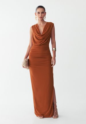 Frau trägt ein rostfarbenes ärmelloses Kleid mit gerafftem Oberteil und drapiertem Ausschnitt, goldenen Schmuck, hält eine geflochtene Clutch und trägt Riemchenabsätze.