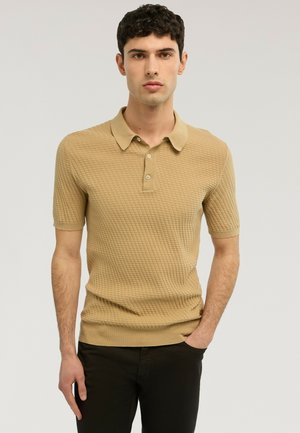 Joven con camisa tipo polo de manga corta de color beige texturizado y pantalones negros, de pie con una mano en el bolsillo frente a un fondo liso.