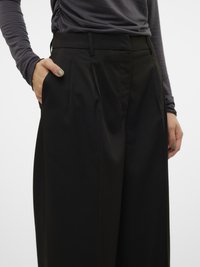 Calças pretas de cintura alta com pregas, apresentando bolsos laterais e textura suave. Combinadas com uma blusa cinza de manga comprida.