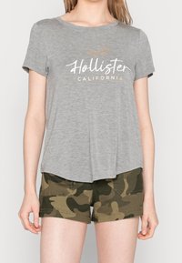 Šedé bavlněné tričko s krátkým rukávem a kulatým lemem, s bílým a zlatým textem "Hollister California". V kombinaci se zelenými maskáčovými šortkami.