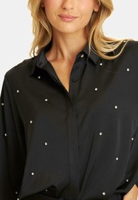 Czarna koszula z kołnierzykiem o gładkiej fakturze, ozdobiona sporadycznymi dodatkami z kryształków i z zapięciem na guziki, częściowo zapięta pod kołnierzykiem.