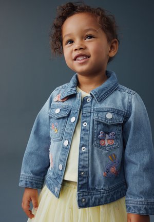 Junges Kind lächelt, trägt eine blaue Jeansjacke mit bestickten Schmetterlingen und Blumen über einem gelben Kleid.